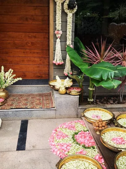 A welcoming entrance for a home-based ceremony, decorated with brass urlis filled with jasmine and rose petals. The traditional rug and hanging floral strings create a warm and inviting atmosphere right from the doorstep.