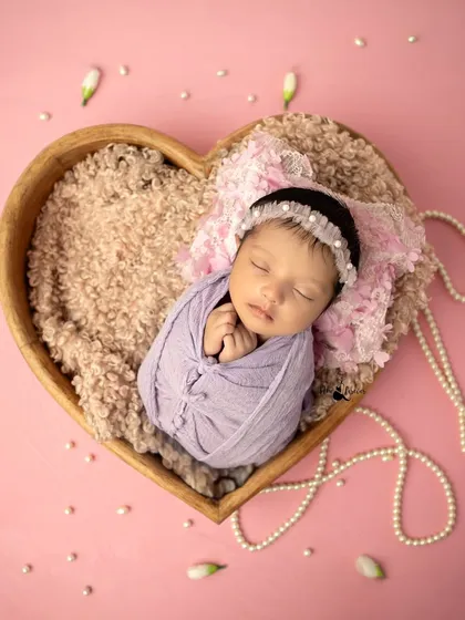 A beautiful overhead shot of a newborn wrapped in a lavender swaddle, resting inside a heart-shaped bowl. The composition is full of love and tenderness.