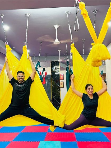 Partner aerial yoga is a great way to connect and build trust. These two students work together to create a beautiful and balanced split pose using the bright yellow hammocks.