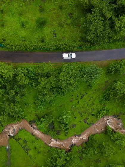 A top-down shot of a car driving on a road next to a winding stream in the outskirts of Mahad, Konkan. The vibrant green of the monsoon creates a stunning contrast.