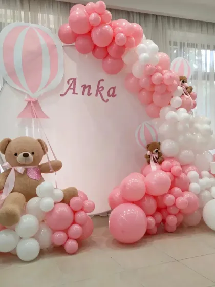 The teddy bear theme is a timeless classic for first birthdays. This setup features a circular backdrop with the baby's name, hot air balloon details, and a beautiful pink and white organic balloon garland.