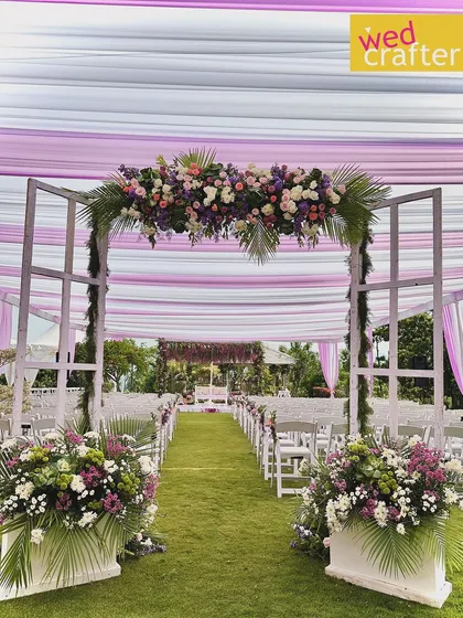 Soothing pastel themes are my jam. This daytime garden wedding features a beautiful aisle entrance framed in white, with lilac and lavender floral arrangements creating a soft, dreamy atmosphere under a draped canopy.