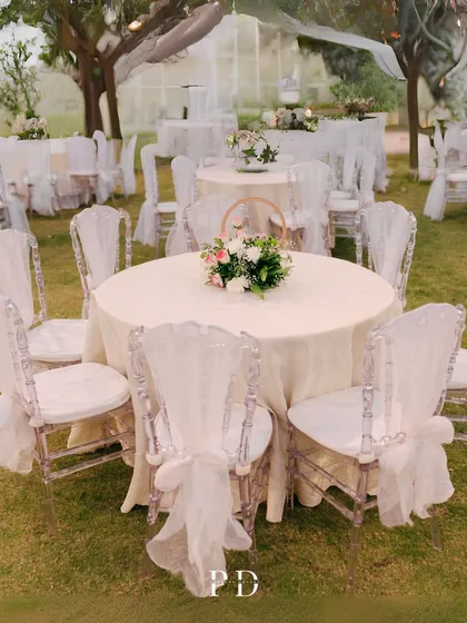 An elegant round-table setup for a reception, using clear princess chairs with delicate white drapes to create a fairytale-like, ethereal feel.