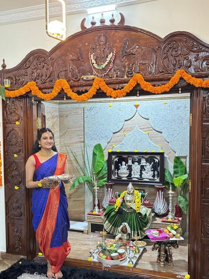 Our client stands before her home's beautifully decorated pooja mandir, ready for the Varamahalakshmi festival in a traditional blue and red silk saree.