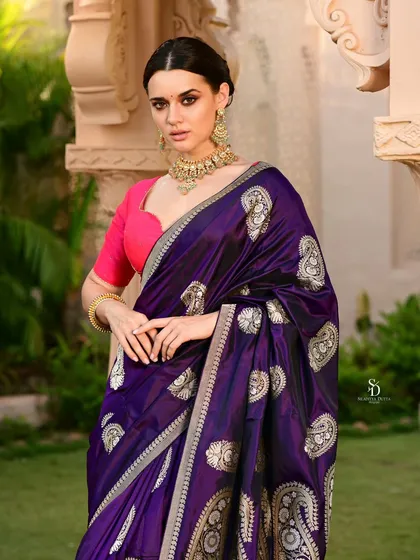 A model wearing a deep purple silk saree with intricate silver motifs. The outdoor garden setting and her poised stance give this traditional look a fresh and regal feel.