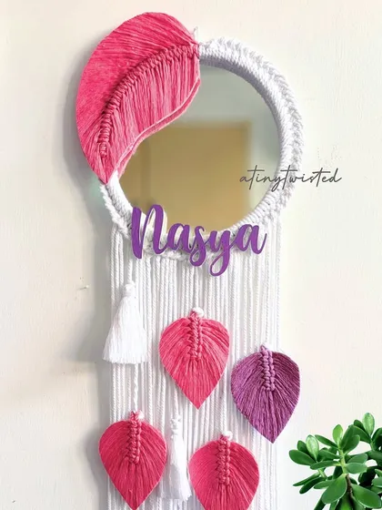 A close-up of a personalized leaf mirror for 'Nasya'. The vibrant pink macramé leaves contrast beautifully with the white-wrapped mirror, making it a stunning and personal piece of decor.