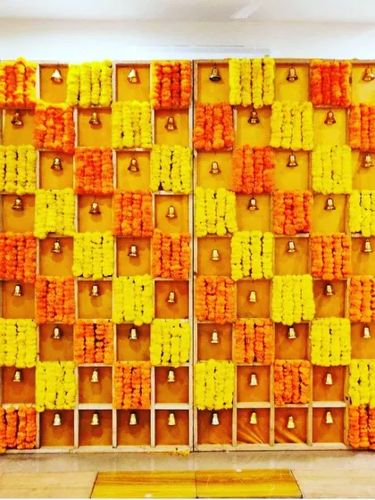 A traditional and festive backdrop crafted from a grid of yellow and orange marigolds, with small brass bells that add a touch of charm and sanctity. This style of decor is perfect for Haldi ceremonies, poojas, or creating a vibrant photo booth.