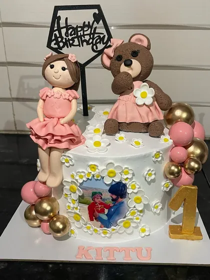 This first birthday cake is full of personal touches, featuring handcrafted fondant figures of a little girl and her teddy bear. It also includes a small, edible photo, combining a fun theme with a cherished memory.