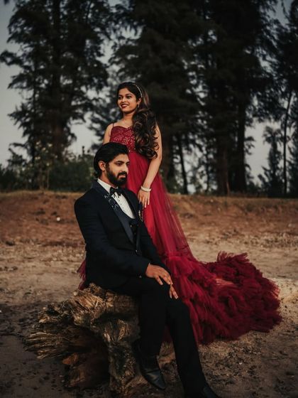 A couple posing during their pre-wedding shoot in the woods. The man is seated on a log while the woman stands, showcasing her beautiful red trail gown.