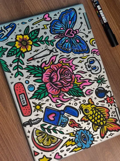 A full-color doodle-style collage painted on a laptop cover. It features a mix of traditional tattoo art, quirky symbols, and vibrant colors.