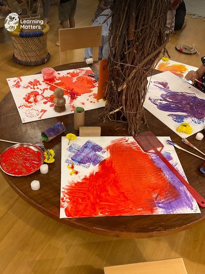 This table is a canvas for collaborative art. Using various tools like rollers and fly swatters, children explore different ways to apply paint, discovering new patterns and textures together. The focus is on shared creation, not a single outcome.