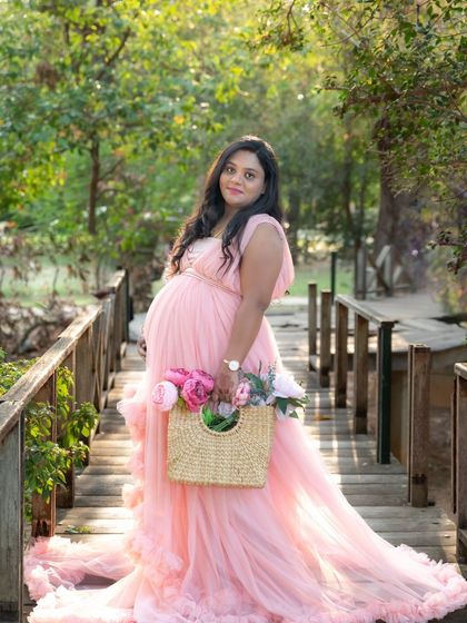 A lovely portrait of a mom-to-be in a pink gown, standing on a wooden bridge in a park. She's holding a cute straw bag with flowers, adding to the charm.