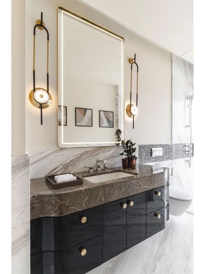 The bathroom continues the modern Art Deco theme with elegant fixtures and luxurious materials. A large, backlit mirror is flanked by stylish sconces, set above a dark vanity with brass knobs.