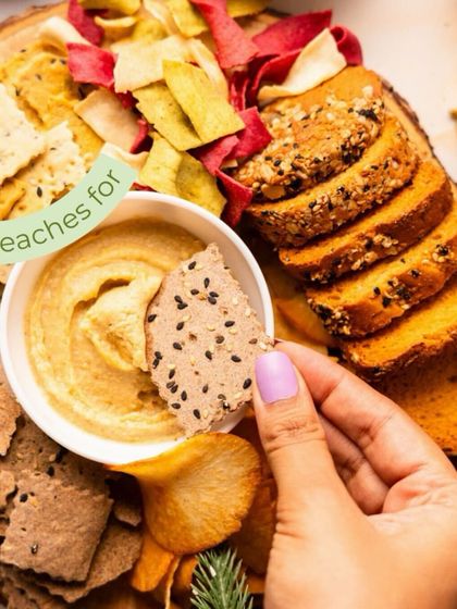 A close-up of a hand dipping a cracker into hummus from a festive platter. Every bite feels like a celebration with my artisanal chips and toasts.