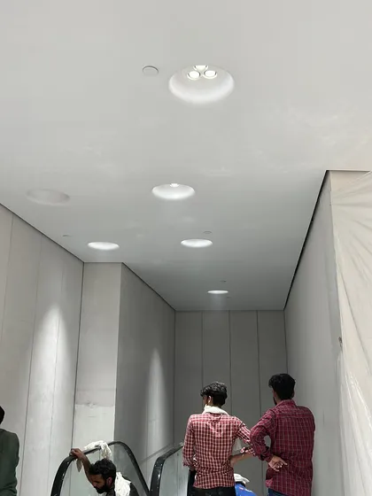 Workers completing the ceiling and lighting installation around the escalator area. The precise placement of recessed lighting is critical for achieving the clean, minimalist aesthetic required in modern retail design.