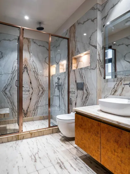 This bathroom showcases stunning book-matched marble with dramatic veining. The rose-gold frame of the shower and the textured wooden vanity add warmth and a touch of color to the luxurious space.