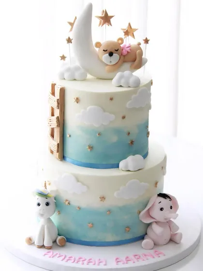 A dreamy cake for twin girls' 1st birthday. This two-tiered design features a teddy bear sleeping on the moon, a unicorn, and a little pink elephant under a starry sky.