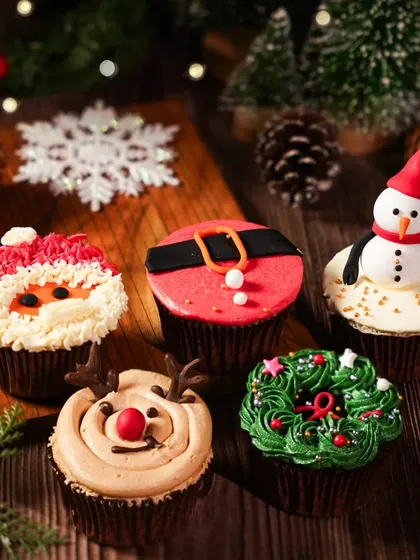 A close-up of assorted Christmas-themed cupcakes, including Santa, reindeer, and wreath designs. The moody, dark wood background makes the colorful frosting stand out.