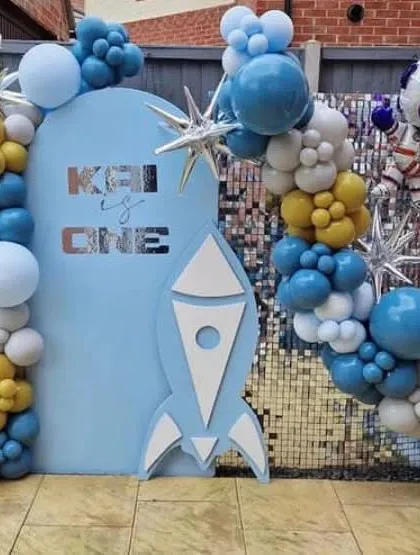 An outer space themed first birthday party on a patio. This decoration features a blue arched backdrop with a rocket cutout, a sequin wall, and a balloon garland.