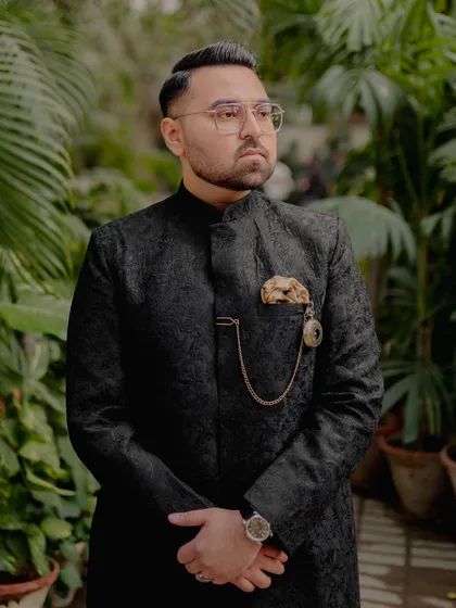 The groom, looking sharp and sophisticated. This portrait captures his distinguished look in a textured black sherwani, complete with a pocket watch, exuding timeless style on his wedding day.