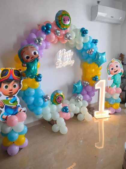 A fun Cocomelon-themed first birthday party. This setup features a circular balloon arch in pastel colors, character cutouts, a 'Happy Birthday' neon sign, and a light-up number '1'.