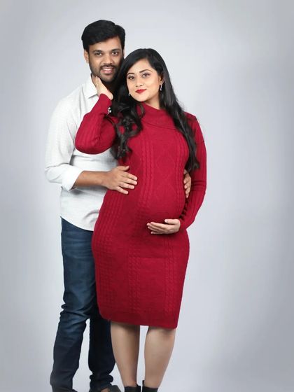 A classic couple's pose that beautifully highlights the baby bump. The mom-to-be looks stunning in her red cable-knit dress, perfect for a cozy studio session.