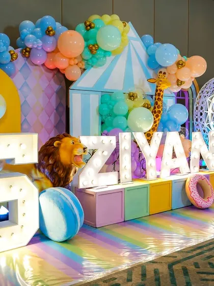A vibrant pastel carnival stage with large marquee letters spelling out the birthday boy's name, surrounded by animal props and a light-up ferris wheel.