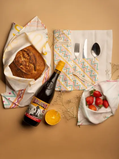 A flat-lay composition showing a variety of fabric accessories in use. This "essentials" shot tells a story of a day out, featuring bread, wine, and strawberries, all wrapped in the brand's products.