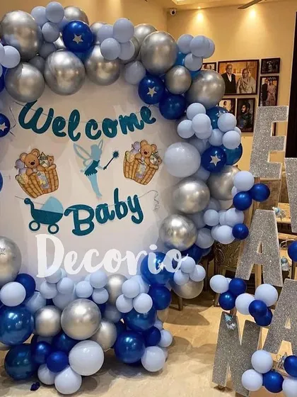 This grand "Welcome Baby" decoration features a large circular backdrop surrounded by a full arch of blue and silver balloons. The sparkling marquee letters spelling out the baby's name add a personalized and glamorous touch.
