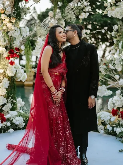 A candid moment of love as the couple stands before a lush floral arch. The bride's red lehenga beautifully contrasts with the white and green floral decor, showcasing how my designs serve as the perfect backdrop for capturing precious wedding memories.