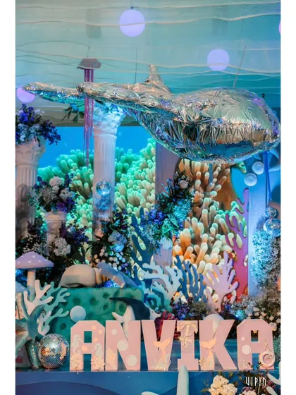 A personalized name sign for 'ANVIKA' set against a vibrant coral reef backdrop with a foil whale balloon floating above. This creates a fantastic focal point for photos.