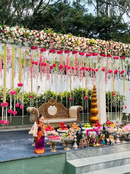 An exquisite outdoor Seemantham mandapam decorated with hanging floral garlands. The stage is beautifully set with an ornate sofa and a traditional display of fruits and sweets.