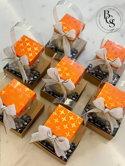 A closer look at these bright orange return gifts. The clear box with a handle is both practical and visually appealing, making it a great choice for festive occasions like Ganesh Chaturthi or a Haldi ceremony.