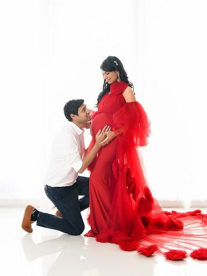 A classic maternity pose with the dad-to-be kneeling to talk to the baby. The simple white background and the flowing red ruffled gown create a timeless and emotional portrait.