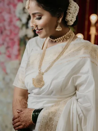 A close-up of the bride's portrait, focusing on her serene expression and the exquisite details of her traditional gold jewelry and white wedding saree.