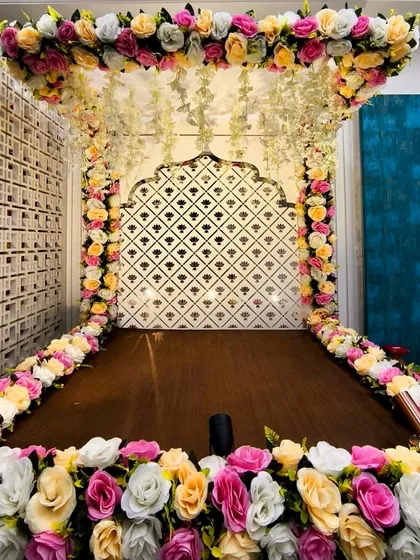 A beautiful Ganpati mandap designed for a home celebration. The structure is decorated with dense floral arrangements in pink, yellow, and white, with hanging jasmine strings for a traditional touch.