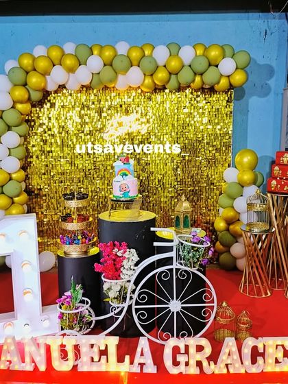 A first birthday setup featuring a gold shimmer wall with a green and white balloon arch. The decor includes a decorative bicycle and marquee letters for the name.