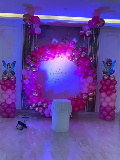 A beautiful "Welcome Our Little Princess" setup in a living room, illuminated with purple lighting. The pink and gold balloon ring creates a glamorous photo backdrop for your home party.