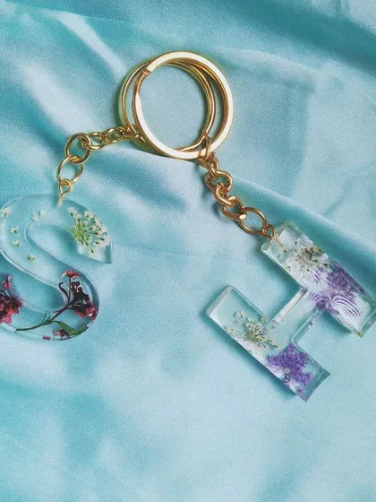 Custom initial keychains made with different types of preserved flowers. The 'S' has delicate red flowers, while the 'H' has small purple and white blossoms.