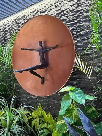 A closer view of the dancing figure, captured in a moment of joyful movement. The warm copper of the sculpture contrasts beautifully with the cool tones of the stone and the vibrant greens of the plants.