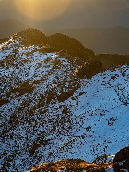 A drone photograph capturing the golden light of the sun hitting a snow-dusted peak in Uttarakhand. This image highlights the textures of the rock and snow, showing the raw beauty of the mountains during the golden hour.