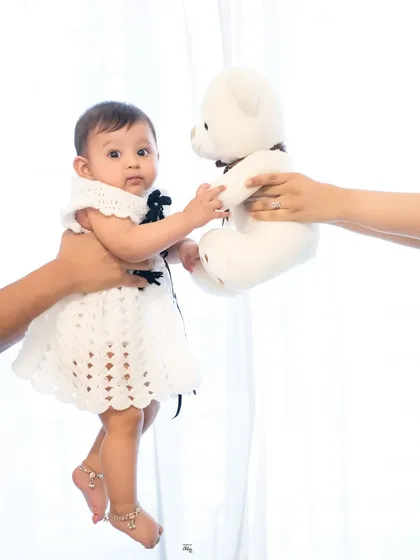 A creative and sweet portrait against a bright white background. Parent's hands hold the baby and a teddy bear, creating a playful interaction and a memory of how small she once was.