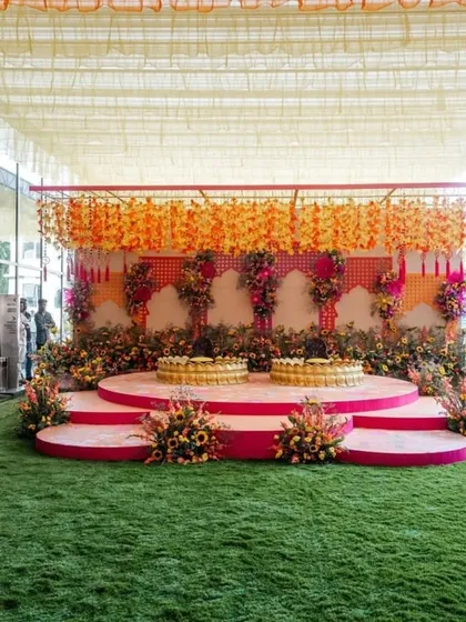 The main stage for a haldi ceremony. The tiered pink stage features two large golden lotus-shaped seats for the couple, set against a backdrop of hanging flowers.