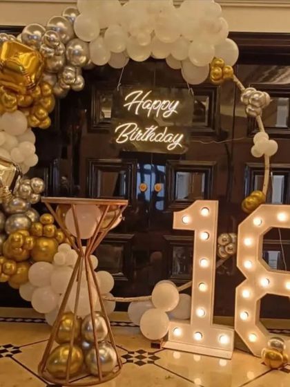 A grand 18th birthday celebration with a gold, white, and silver theme. This setup features a balloon arch against a glossy black door, a neon sign, and oversized light-up marquee numbers for '18'.
