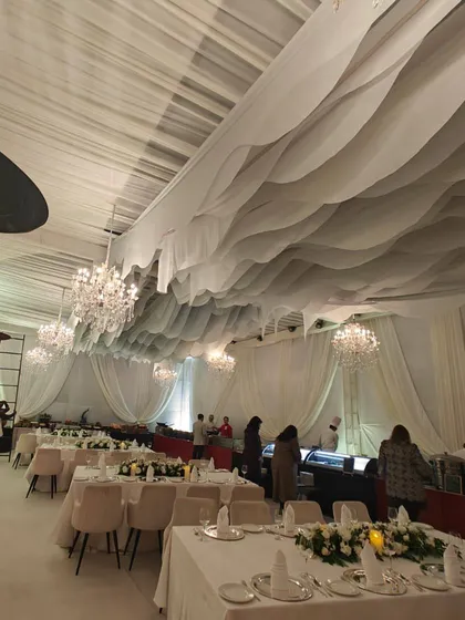 A wider angle of the all-white reception dining area, showing the full effect of the wavy ceiling drapes and multiple crystal chandeliers that create a sense of movement and elegance.
