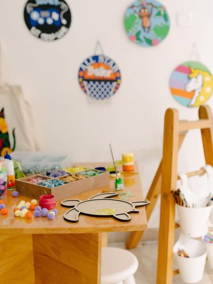 This is a typical setup for one of our craft sessions. I make sure all the materials, from colorful pom-poms and mosaic tiles to paints and wooden bases, are neatly organized and ready for our artists to start creating.