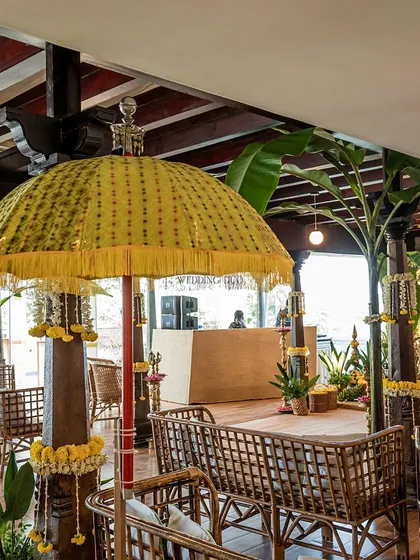 A traditional, ornate yellow parasol provides shade and a pop of color over the cane seating at this indoor Haldi ceremony, with the DJ booth visible in the background.