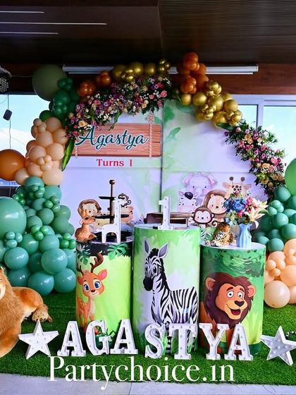 A safari-themed decoration set up on a balcony. This arrangement features a lush green and gold balloon arch, a personalized backdrop, and plush animal toys like a lion and tiger to create a cozy jungle corner.