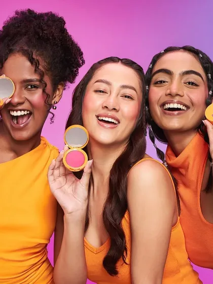 A bright and happy campaign image featuring models with colorful blush compacts. The makeup is radiant and playful, with an emphasis on healthy, flushed cheeks and big smiles.