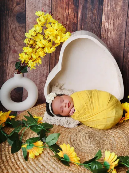 A heart-shaped prop filled with sunshine. This newborn is wrapped in bright yellow, sleeping peacefully in a heart bowl next to a vase of sunflowers.